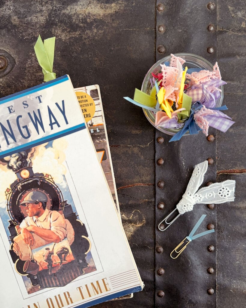 A green ribbon paperclip bookmark in Ernest Hemingway's book, "In Our Time," with a glass jar full of bookmarks sticking out and a couple strewn across the table