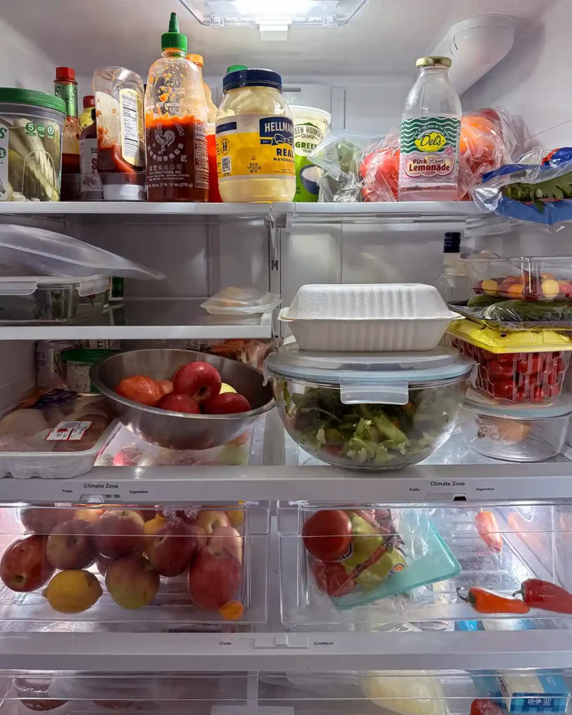 The interior of a refrigerator with cluttered and messy shelves full of vegetables, fruits, condiments, and much more