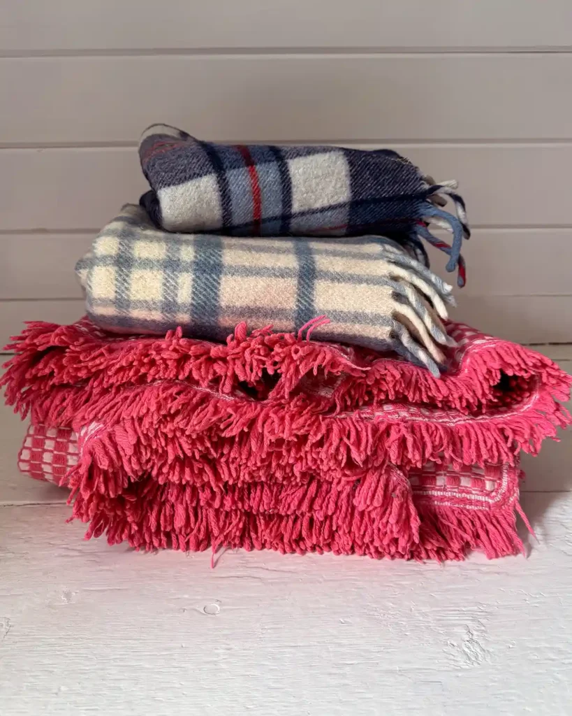 A stack of vintage blankets purchased from the Brimfield flea market. On the bottom, a woven hot pink and white coverlet with lots of pink fringe. In the middle a cream and light blue plaid, wool stadium blanket with fringe. And on the top a dark blue, cream, and red plaid, wool stadium blanket with fringe.