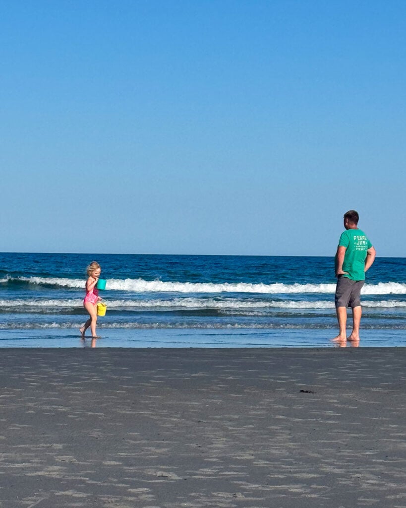A small kid with sand buckets and her Dad on the beach at the edge of the water with a bright blue sky. Beach is York Beach in Maine.