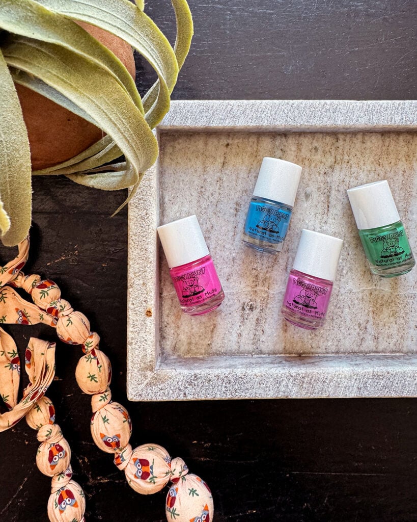 Four shades of bright Piggy Paint nail polish (pink, blue, purple, and green) sitting in a marble tray on top of a black desk by a plant and fabric bead necklace