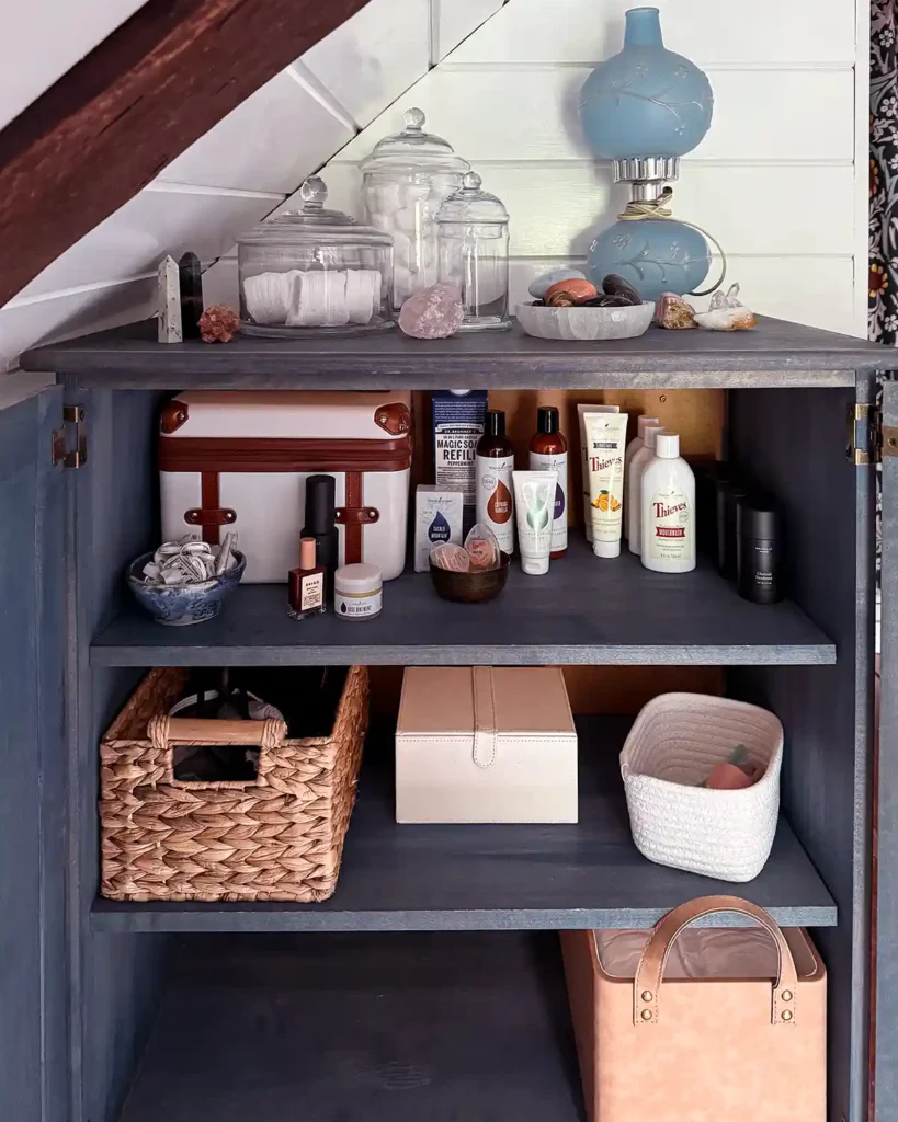 A blue bathroom cabinet decluttered and organized with empty space next to a faux leather basket on the bottom shelf. Extra bath products, lotions, nail polish, etc. on the top shelf. A basket of hair tools, jewelery box, and pedicure tools on the middle shelf.