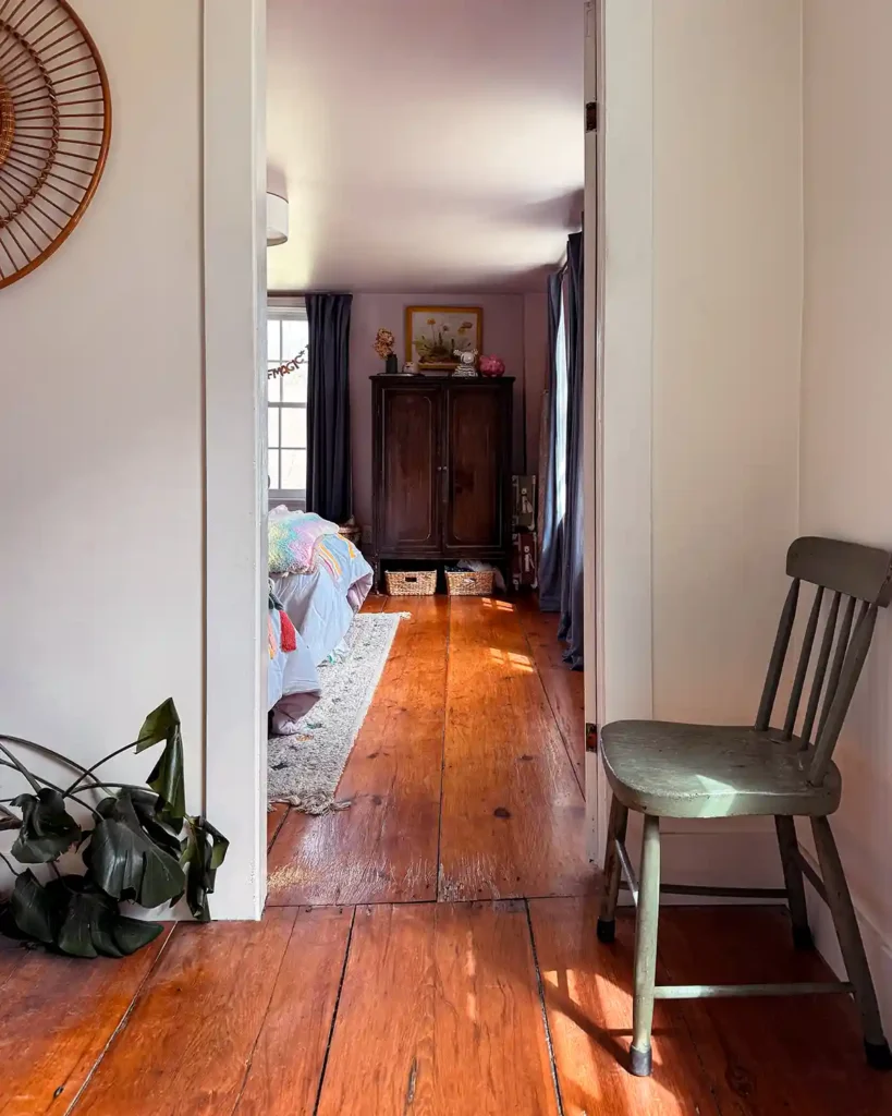 A girls' shared bedroom color-drenched a light pink color with both the walls and the ceiling painted to match. View of the room from the doorway with a chair and plant sitting outside the door. Inside the room is a vintage wardrobe and pair of antique twin beds with a rainbow comforter
