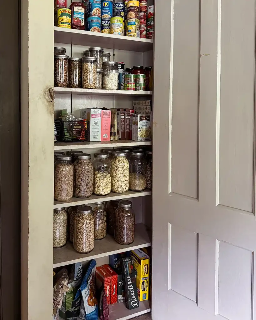 A kitchen pantry cabinet (a vintage jelly cabinet) decluttered and organized featuring large glass jars of pantry ingredients, snacks, canned goods, extra condiments, nuts, cereal, and more