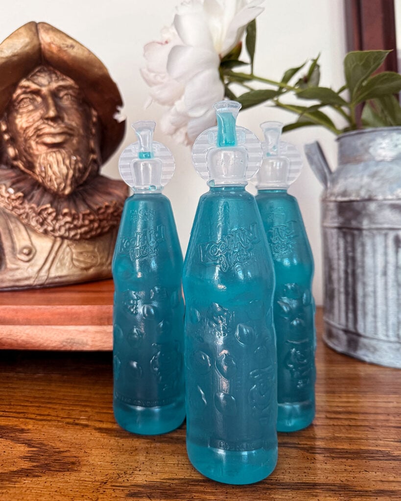 Three bottles of Kool-Aid Bursts sitting on a dining room buffet with a galvanized jug of peonies in the background along with a statue bust
