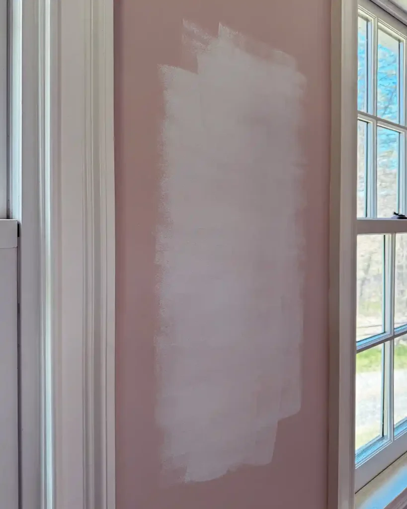 A piece of drywall that was previously painted pink, between two windows that has been patched and primed with a stain-blocking primer before painting the wall pink again