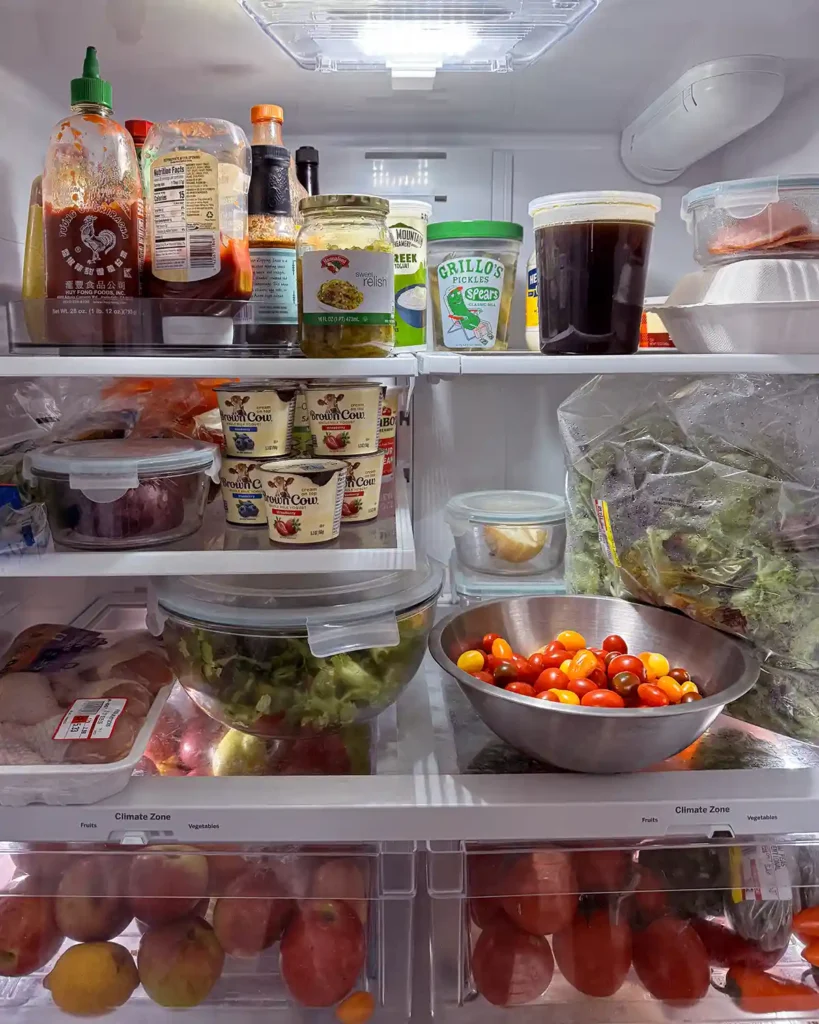 A fully stocked refrigerator after decluttering a grocery shopping with a lazy susan of condiments, bowl of tomatoes, bag of lettuce, drawers of fresh fruits and vegetables, and more