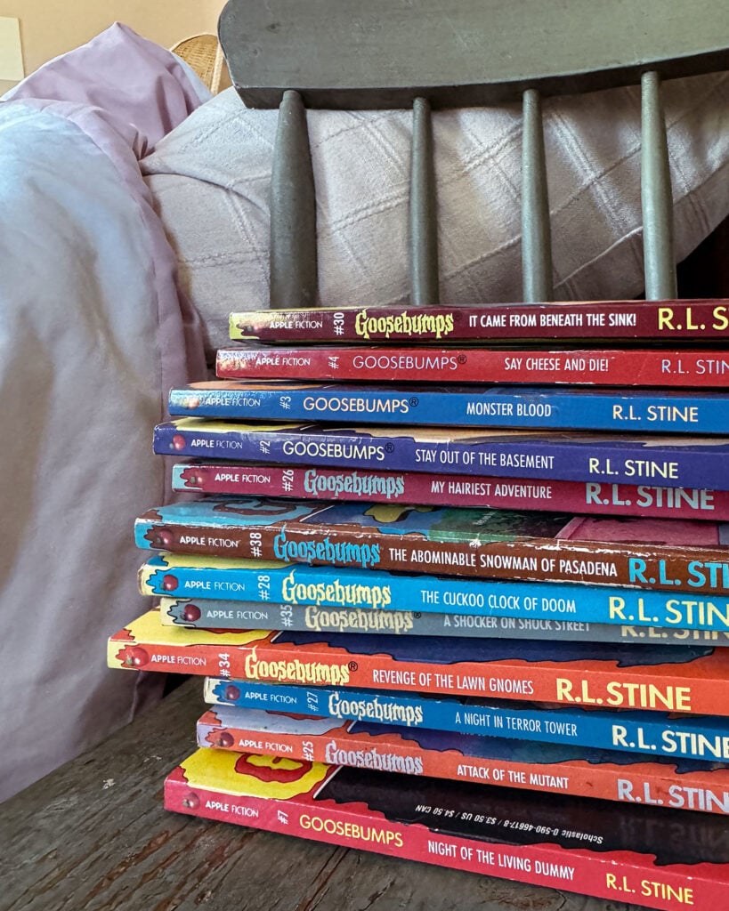 A stack of 12 vintage Goosebumps books sitting on a chipped paint chair beside a bed