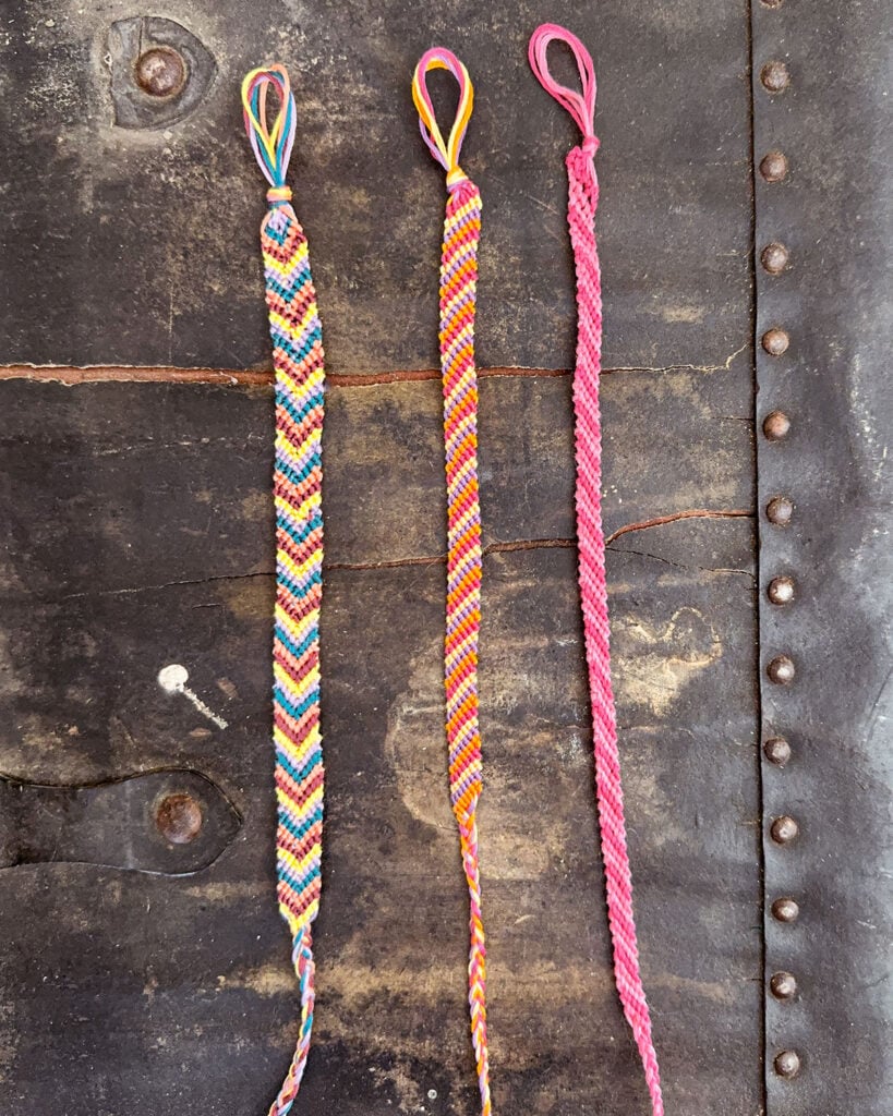 A trio of colorful friendship bracelets using pink, yellow, orange, dark blue, and peach embroidery floss in striped and chevron patterns