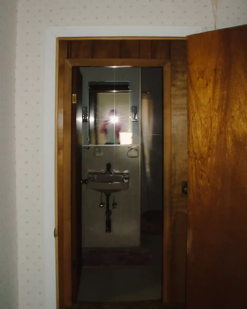 A before shot of a dated bathroom looking into the room from the bedroom across the hallway; bathroom features a frameless mirror, hollow core door, and a light pink wall mounted sink
