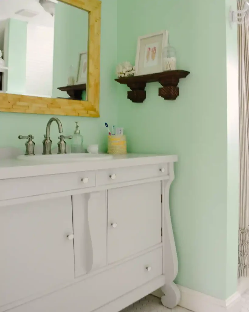 A light gray painted bathroom vanity next to a bathtub/shower combo; wood framed mirror hanging above and a wooden shelf on the wall