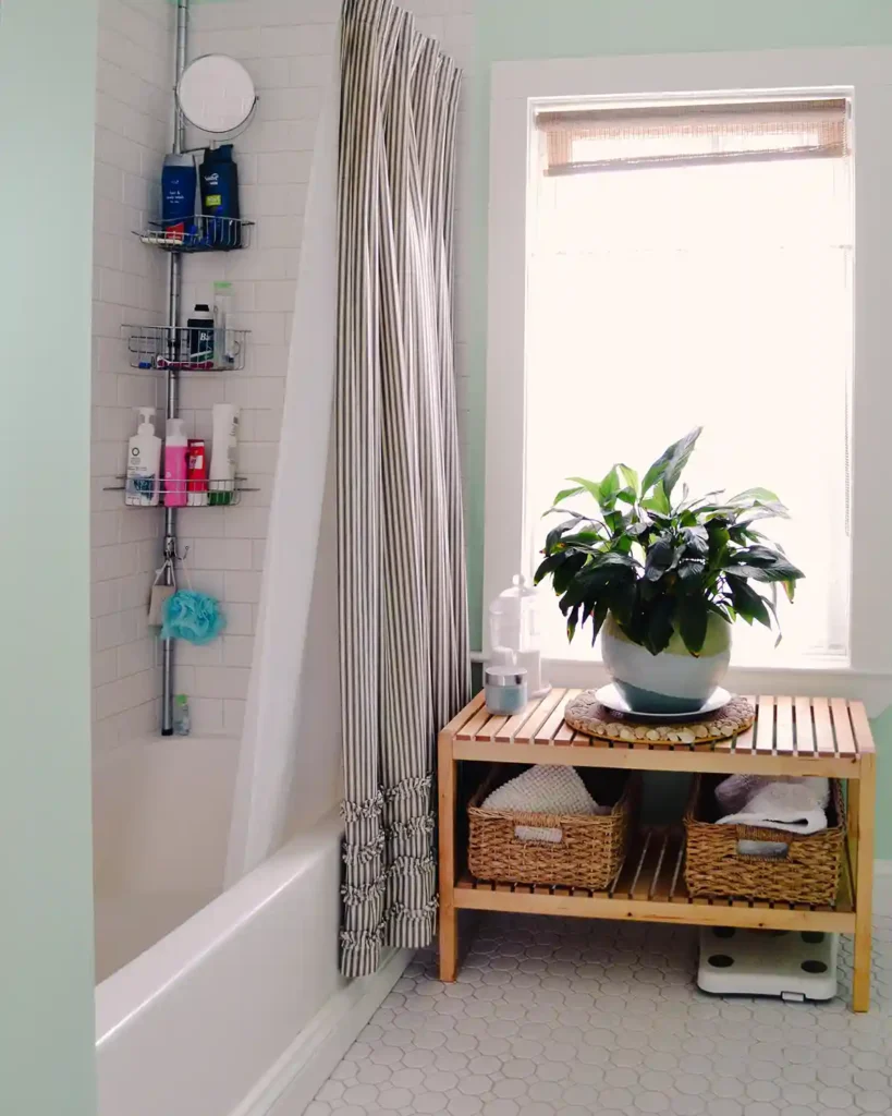 A small, bright, and airy cottage-style bathroom with white hex floor tiles, mint green walls, white subway tiled shower with white grout, a wooden bench in front of a window with a plant, and a corner shower caddy