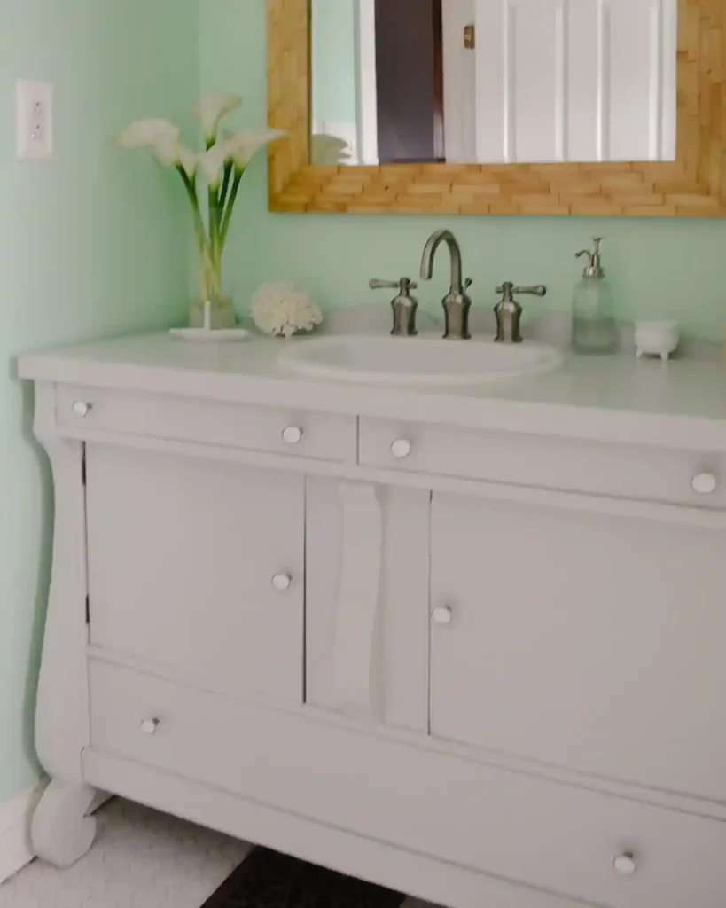 A vintage buffet turned into a light gray painted, cottage-style bathroom vanity with a wood tile framed mirror hanging above the vanity