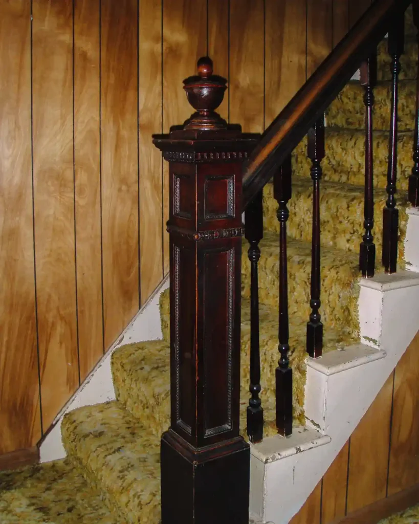 A dated, carpeted staircase with old, shaggy carpeting, dark stained newell post and spindles, broken spindle, faux wood paneling adjacent to a hallway