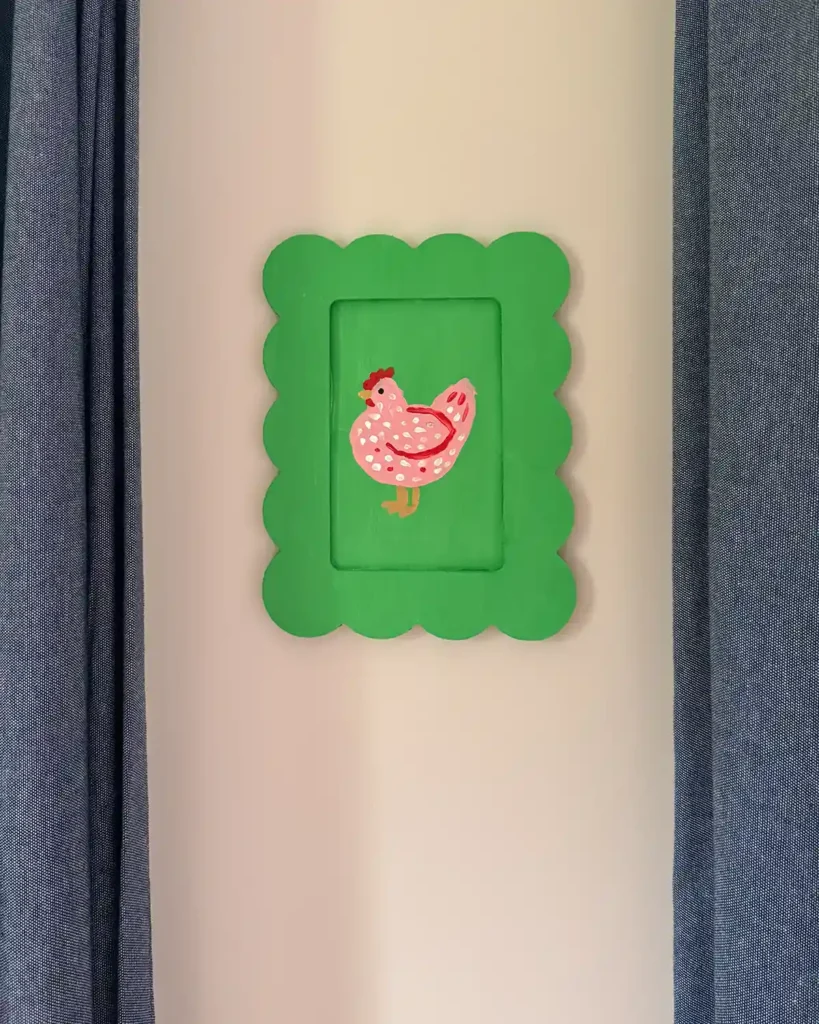 A DIY folk-art inspired chicken print hanging on the wall as kids' bedroom decor; green background with a pink chicken on it hanging on pink walls between two chambray curtain panels