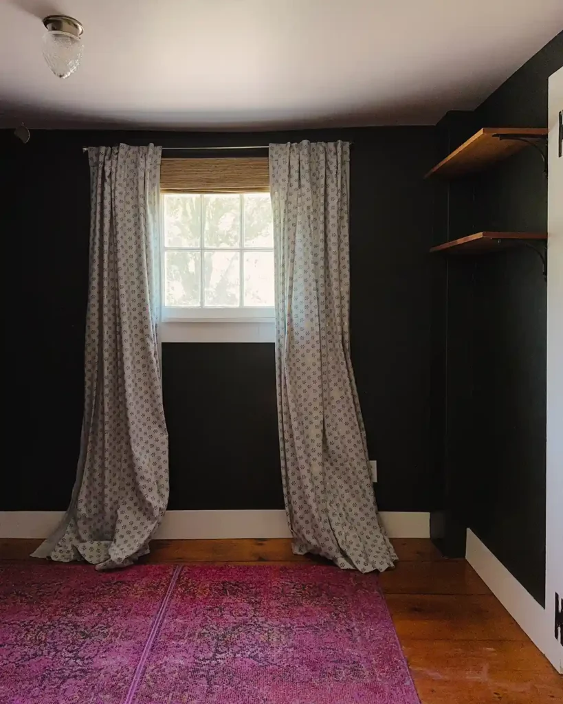 An old nursery that is completely cleared out except for the pink rug, light blue curtains with a woven shade, and some wall shelves
