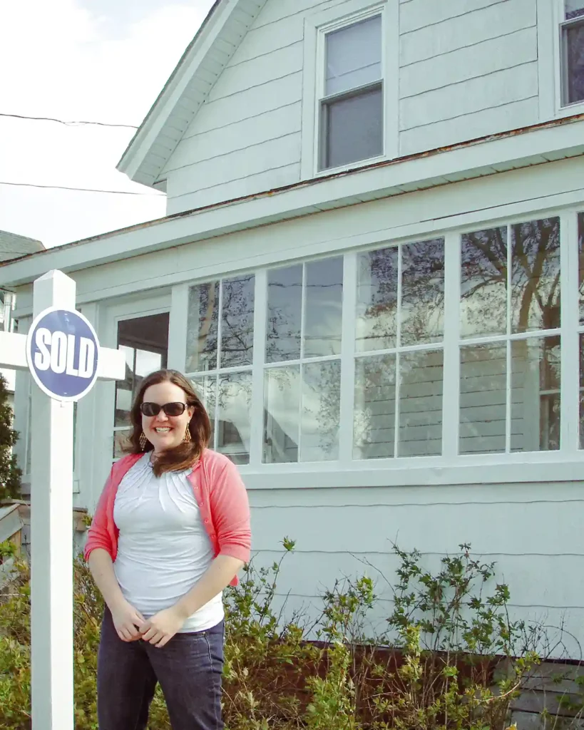 A young woman standing out front of a white farmhouse with an enclosed front porch next to a sold sign on the day she closed on the purchase of her first home