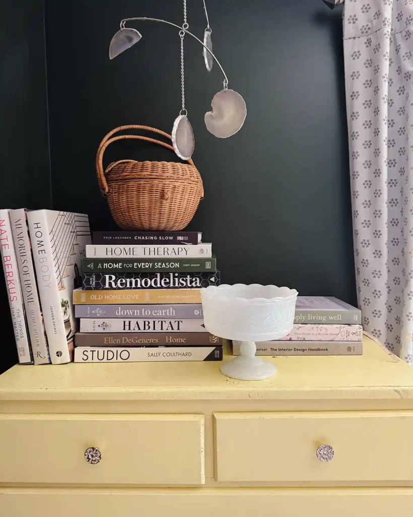A collection of home design books sitting atop a light yellow dresser with an apple shaped basket and milk glass bowl mixed in