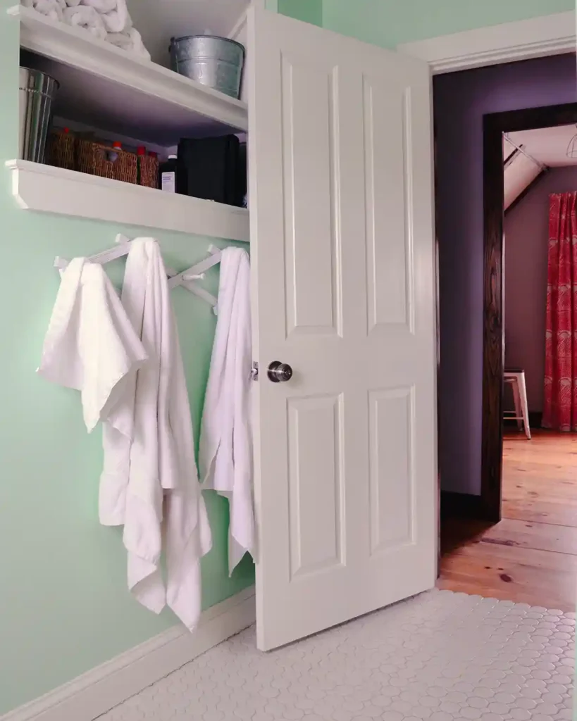 The storage area of a mint and white cottage style bathroom with shelves carved into the empty space in a wall and white towels hanging below; the bathroom's white door is open with a view of the room across the hall peeking through
