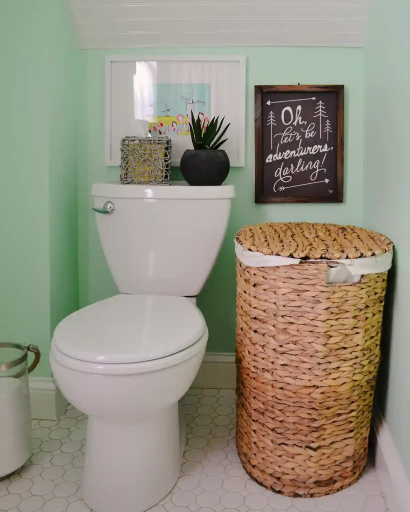 The toilet area in a small, cottage-style bathroom remodel with a white toilet under a slanted beadboard clad ceiling, large woven laundry hamper, art hanging behind the toilet, and a plant and tissue box on the back of the toilet; mint green walls and white hex floor tiles