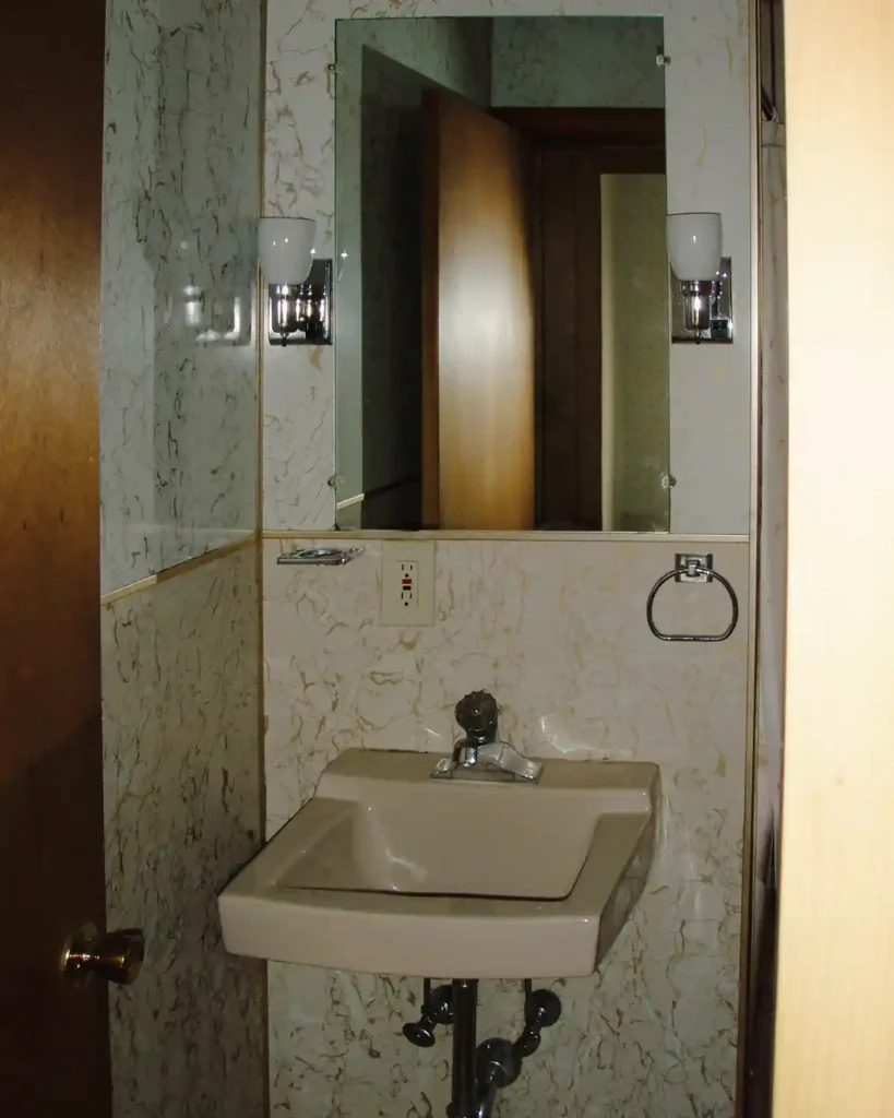 A dated bathroom sink nook before renovating, featuring a pink wall-mounted sink with an unframed mirror and lighting sconces