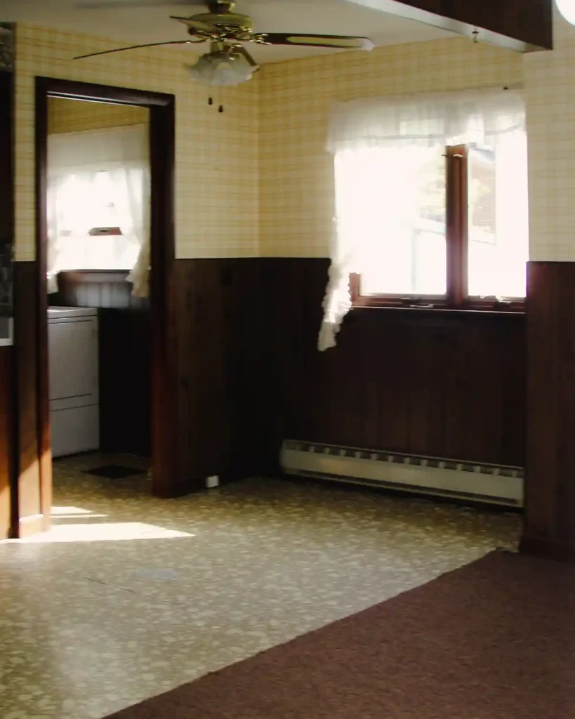The eat-in-kitchen side of a dated kitchen with dark paneling and yellow, plaid wallpaper, old ceiling fan, dated carpet and yellow linoleum flooring, and peek into an old laundry room before renovating