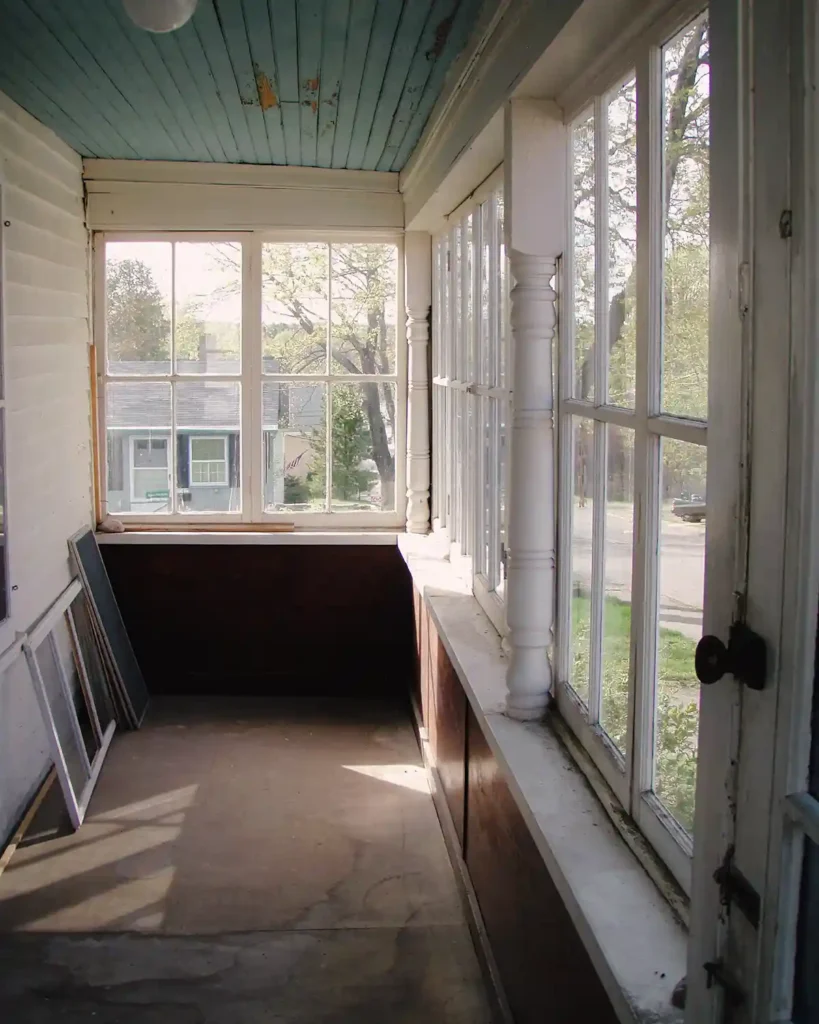 An enclosed front porch with a light blue bead board ceiling, lots of windows and light, screen door, and water damaged subflooring