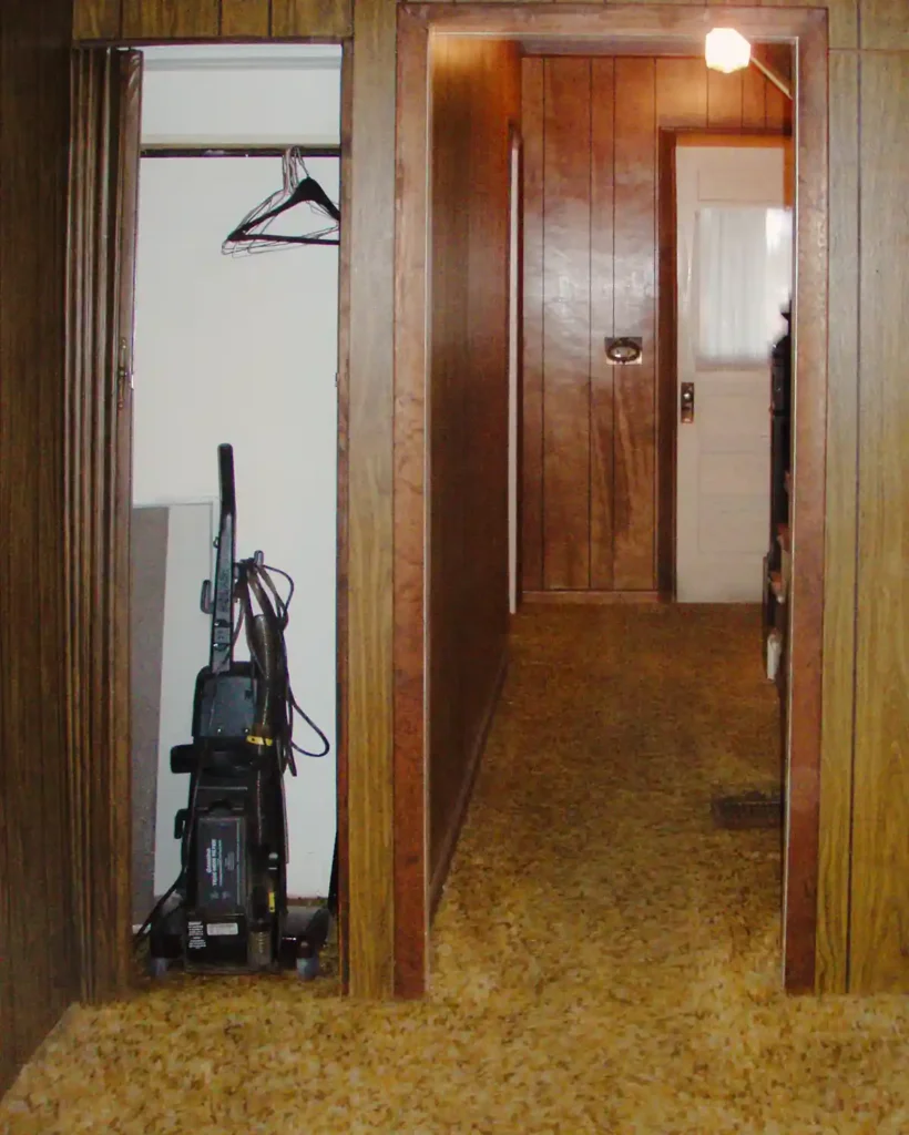 A first floor hallway in a dated home before renovating featuring shaggy brown/yellow rug, a closet with a vacuum, long hallway to a front door, and vintage lighting