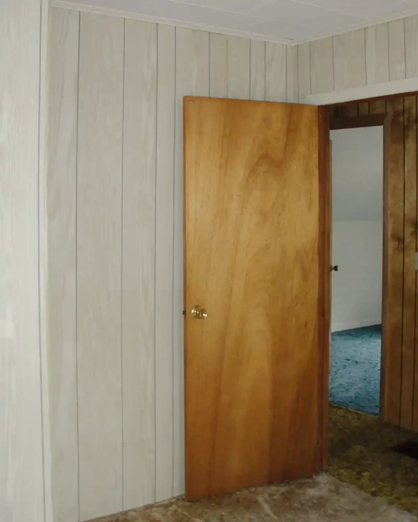 A dated guest bedroom with faux wood paneled walls, brown shag rug, hollow core wood door, and a drop ceiling before beginning a whole house renovation
