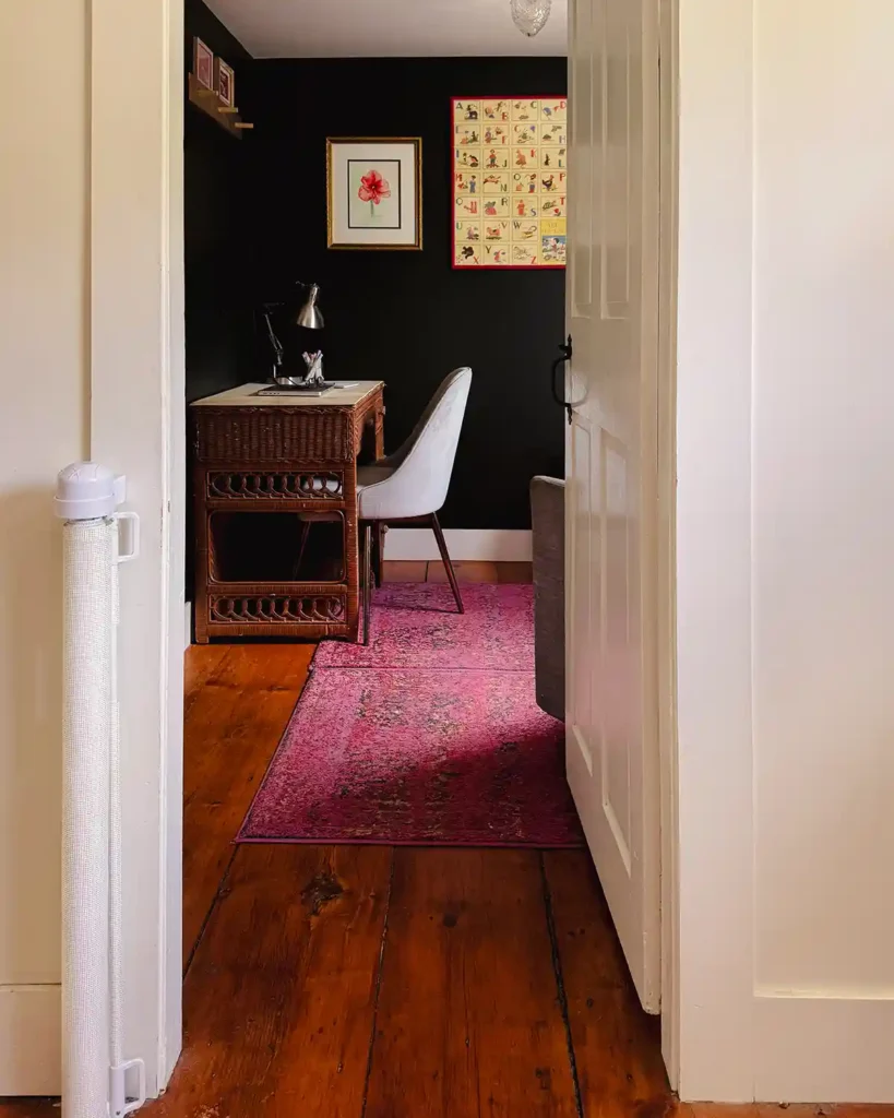 Looking through a doorway into a small, home office with a wicker desk, fabric desk chair, pink rug, and dark green (almost black) painted walls