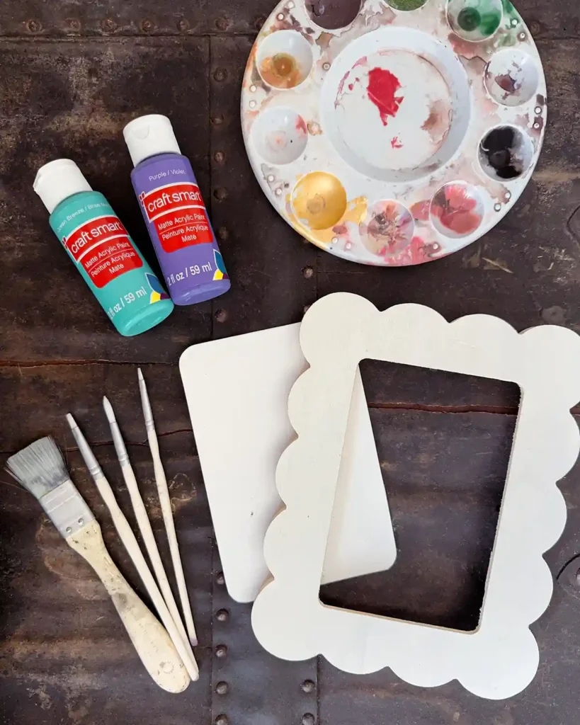 All the tools and supplies for a kids art project including a paint tray, colorful paints, wood frame, watercolor paper, and paint brushes