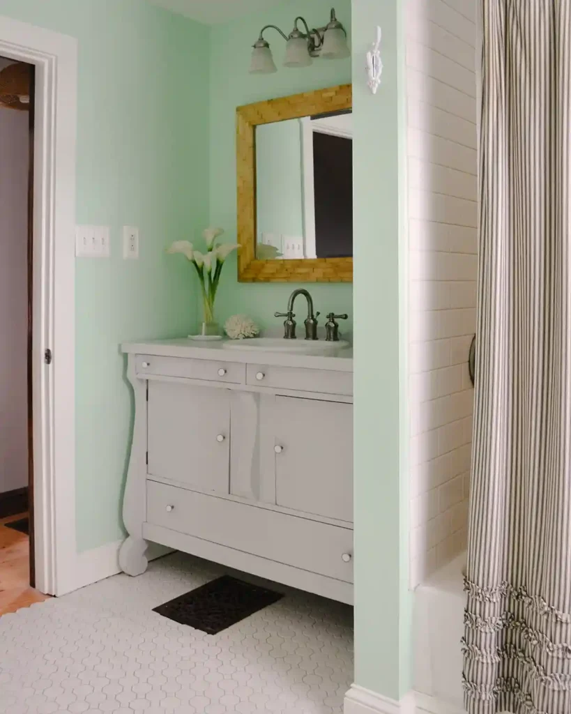 A light gray painted bathroom in a mint-colored painted bathroom with white hex floor tiles; wood framed mirror hanging over the vanity and sink