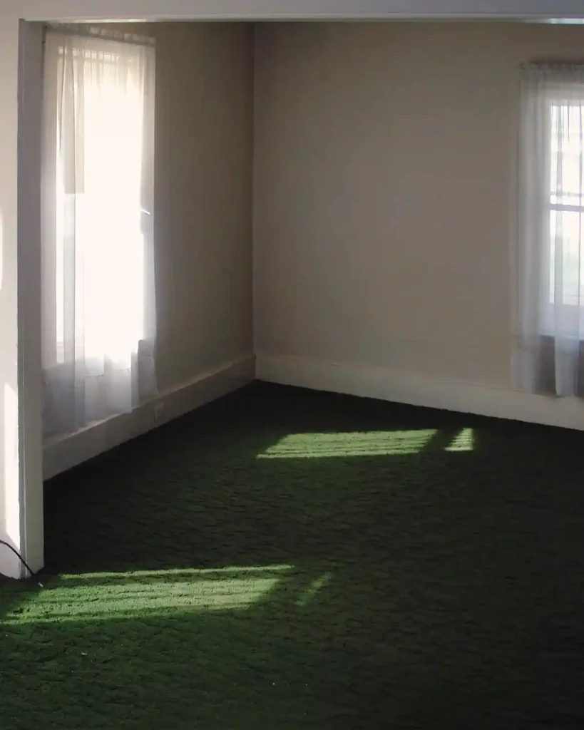 A vintage living room with a shaggy green rug, cream colored walls, and sheer curtains before renovating the space