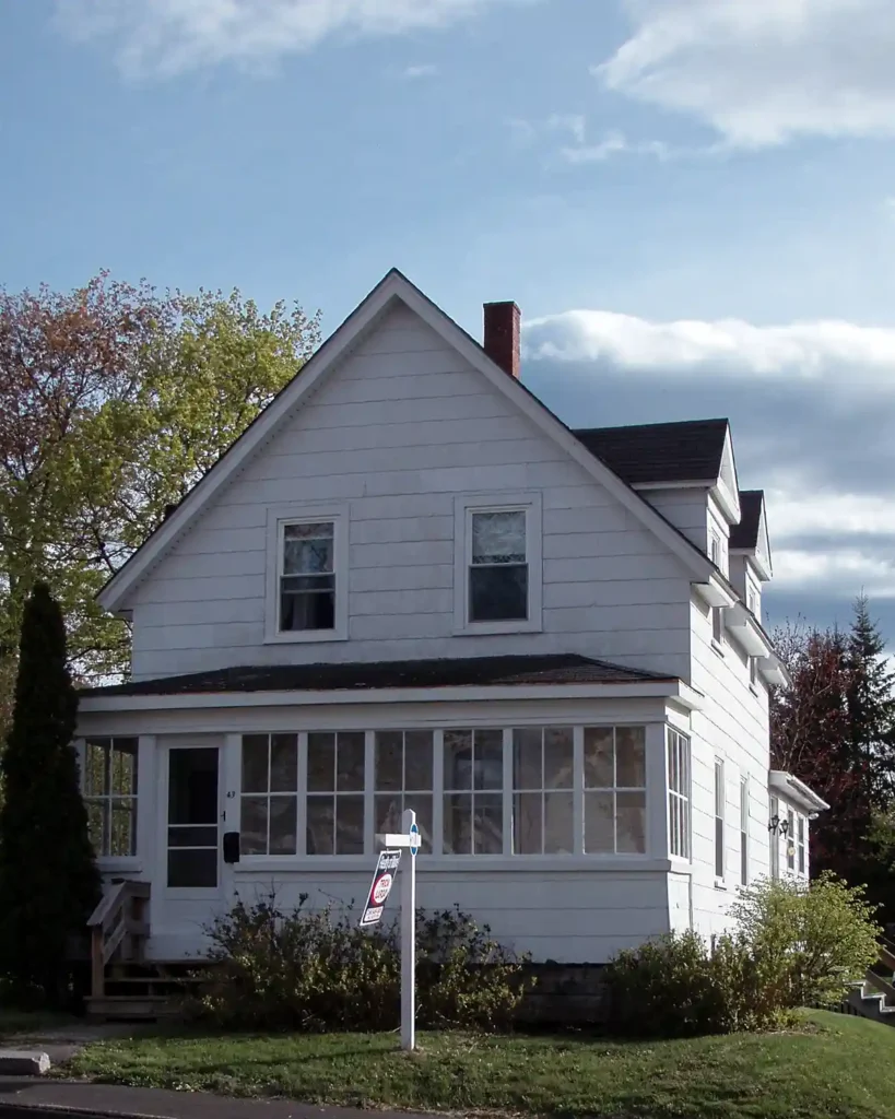 The gable end of a white, Maine farmhouse with enclosed front porch with a for sale sign out front