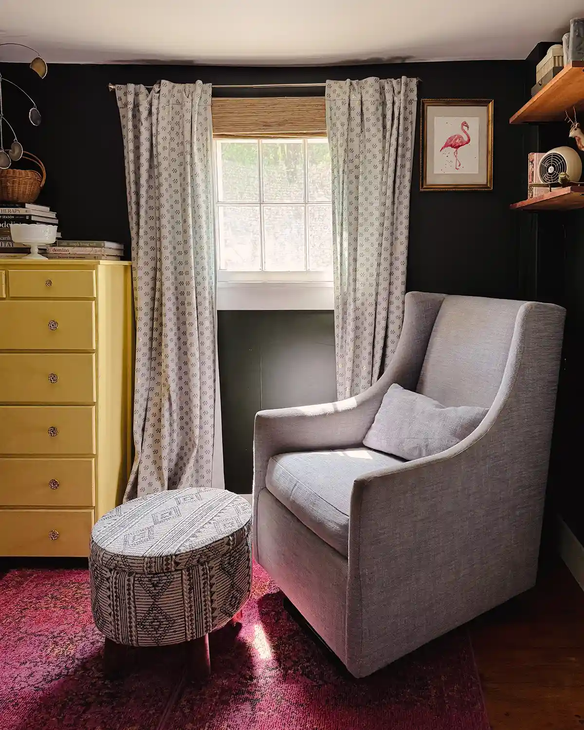 A one day home office makeover featuring a small room with dark walls, light blue curtains, glider rocking chair with a footstool, butter yellow dresser with a stack of books on top, open shelves filled with knick knacks and decor, and a bright pink rug over wide pine floors