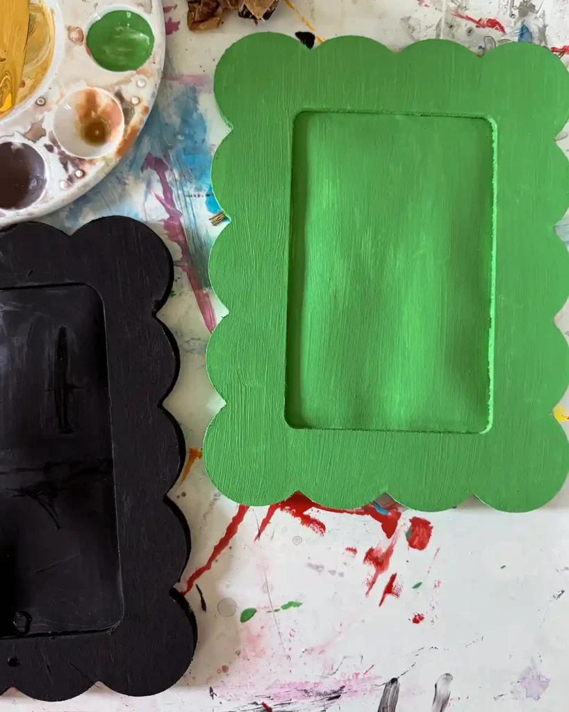 A bright green painted scalloped frame and background sitting beside a black painted one next to a painting tray