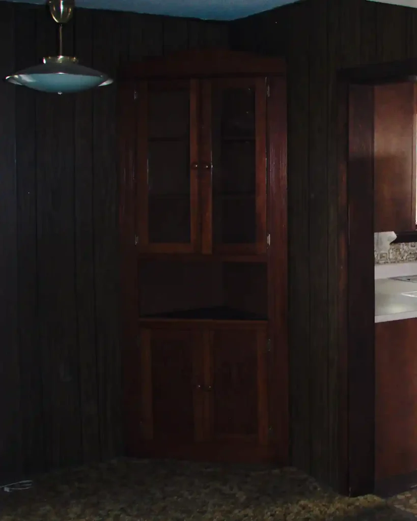 A vintage, dark paneled dining room with dark carpeting and a corner hutch and metal light fixture with a view into the kitchen