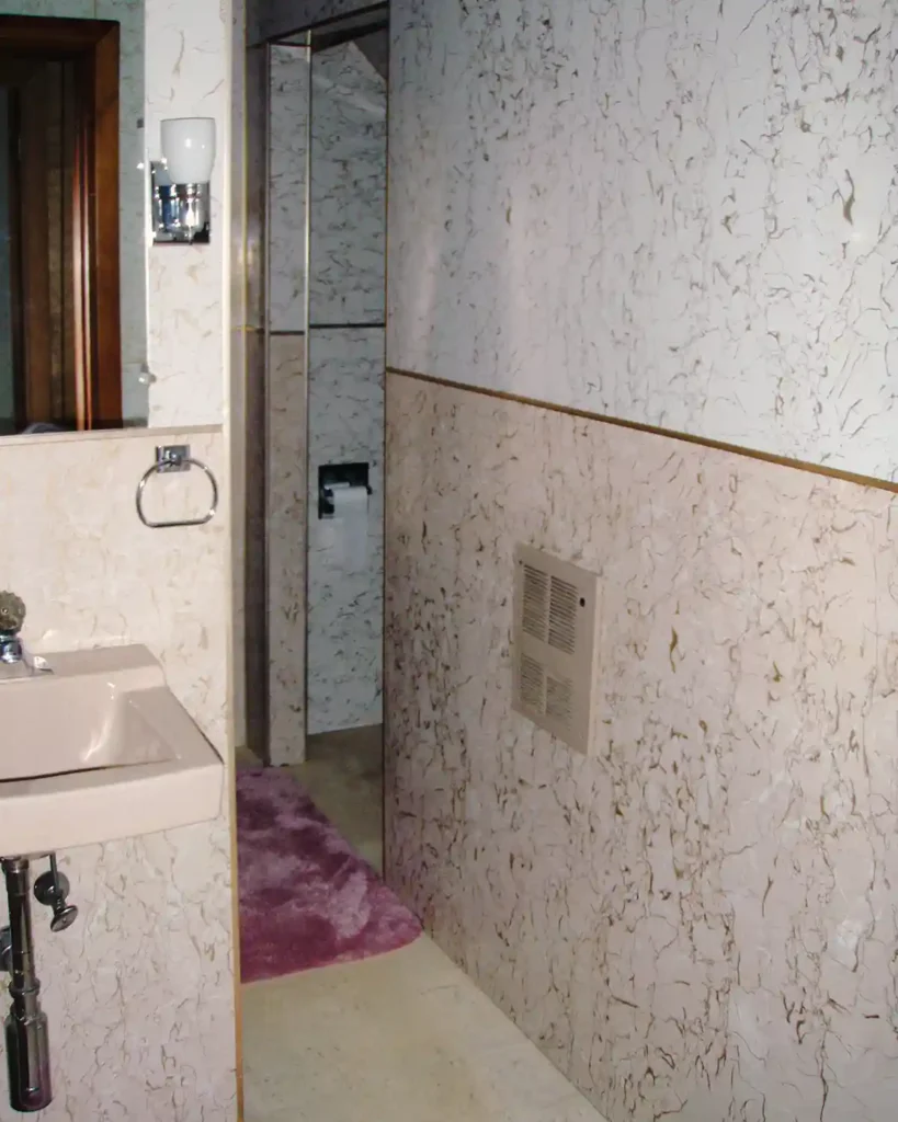 A dated pink paneled bathroom with a toilet nook, pink wall-mounted sink, dated paneling and light fixtures before renovating