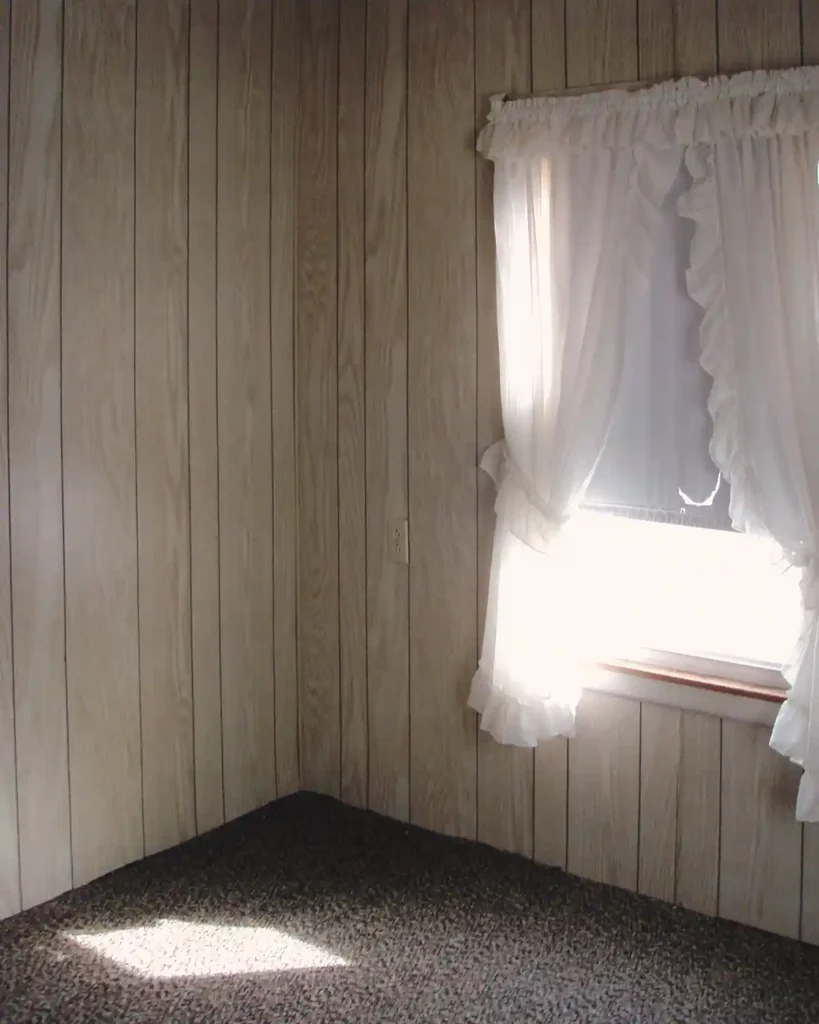 Primary bedroom before remodeling featuring a window with a roller shade and white curtains, light faux wood paneled walls, and a dated flat weave carpet