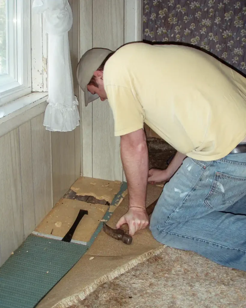 A man kneeling on a floor with a crowbar, pulling up old carpeting and a sub floor in a paneled bedroom checking to see if there are hardwood floors underneath