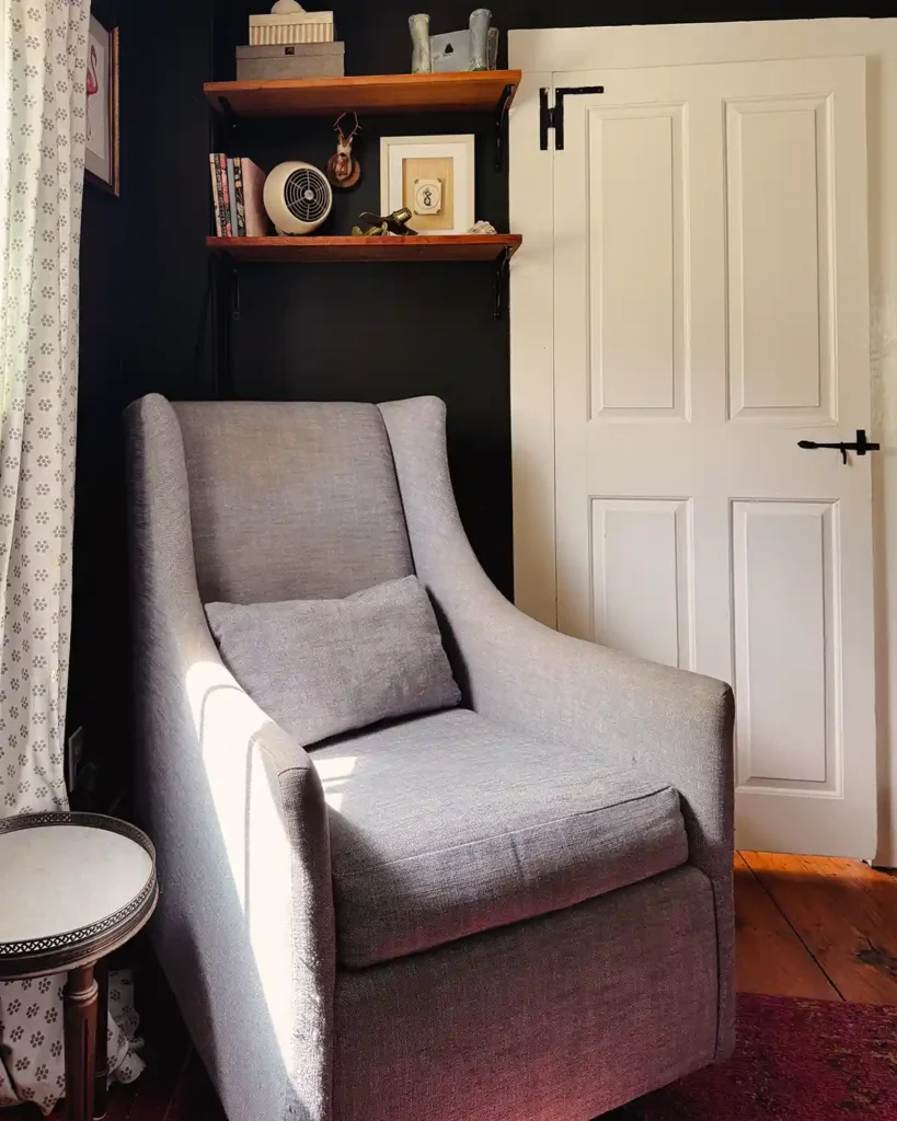 The reading nook in a home office with a light blue gliding chair, marble side table, and shelves filled with journals, notebooks, storage boxes, a small fan, and decor