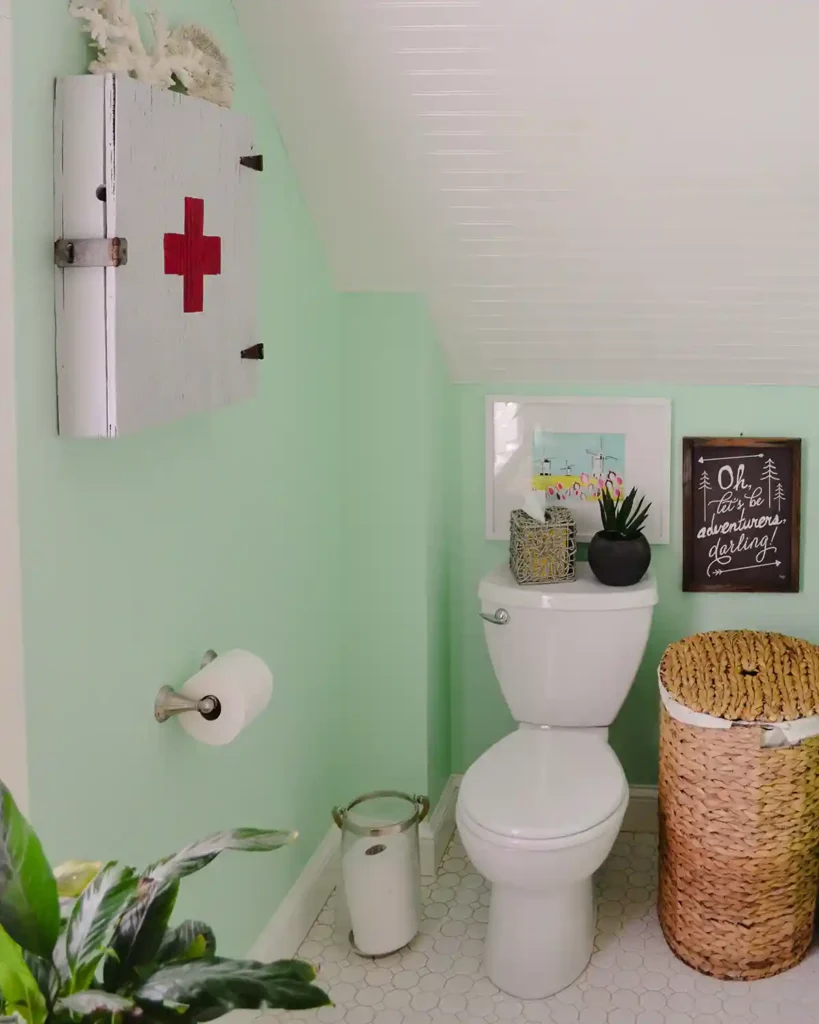 A remodeled cottage bathroom toilet area with a slanted white beadboard ceiling, light mint green walls, toilet next to a laundry hamper, a wooden medicine cabinet hanging on the wall, and a plant peeking out in the foreground