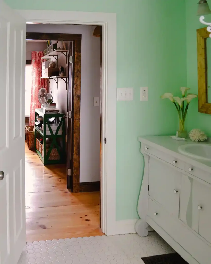 A cottage-style renovated bathroom view from the window looking out the bathroom door and into the room across the hallway; bathroom with light mint green walls, light gray painted bathroom vanity, and white hex tile floors