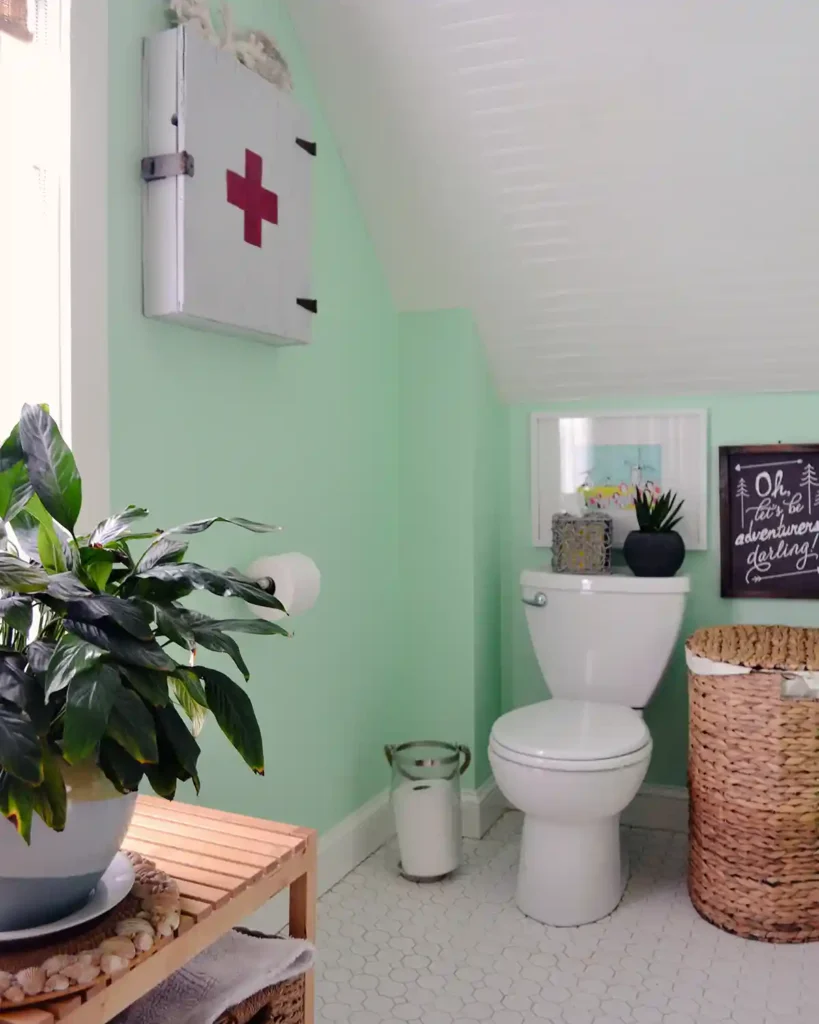 The toilet nook in a small bathroom remodel with a white toilet next to a natural fiber laundry basket under a slanted ceiling with beadboard painted white, walls are minty green, a white first aid cabinet hangs on the wall, and a plant on a wooden bench sits in the foreground