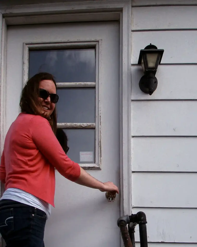 A young woman in a pink cardigan and sunglasses unlocking the white front door to her first purchased home