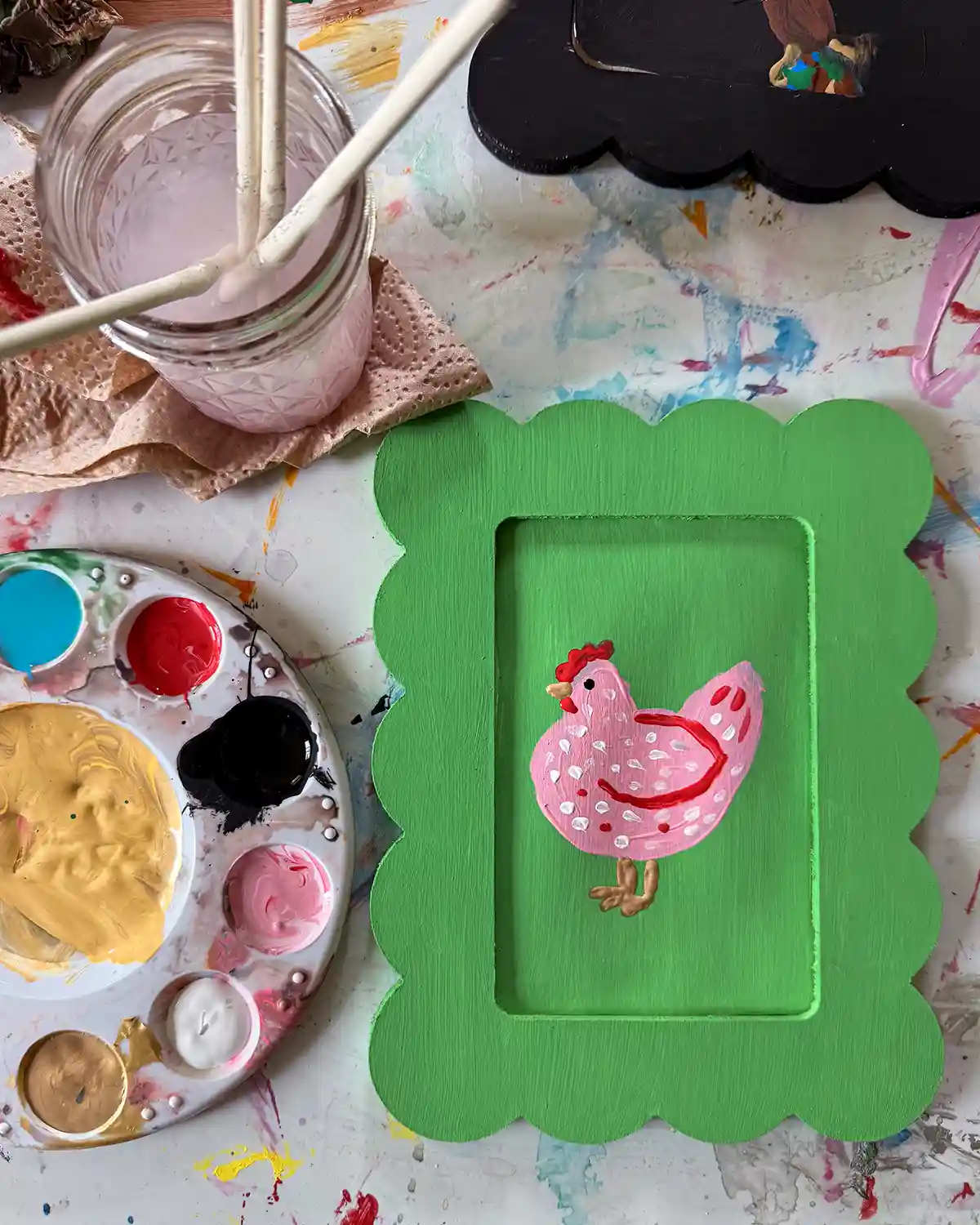 A DIY whimsical chicken art project with both the scalloped frame and background of the print painted bright green with a light pink chicken; surrounded by a paint tray, Ball jar filled with dirty paint brushes, and a black frame
