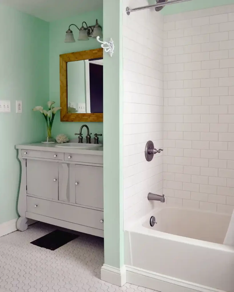 A white subway tile shower with white grout and a white tub sitting next to a light gray bathroom vanity; mint green walls with white hex tiles