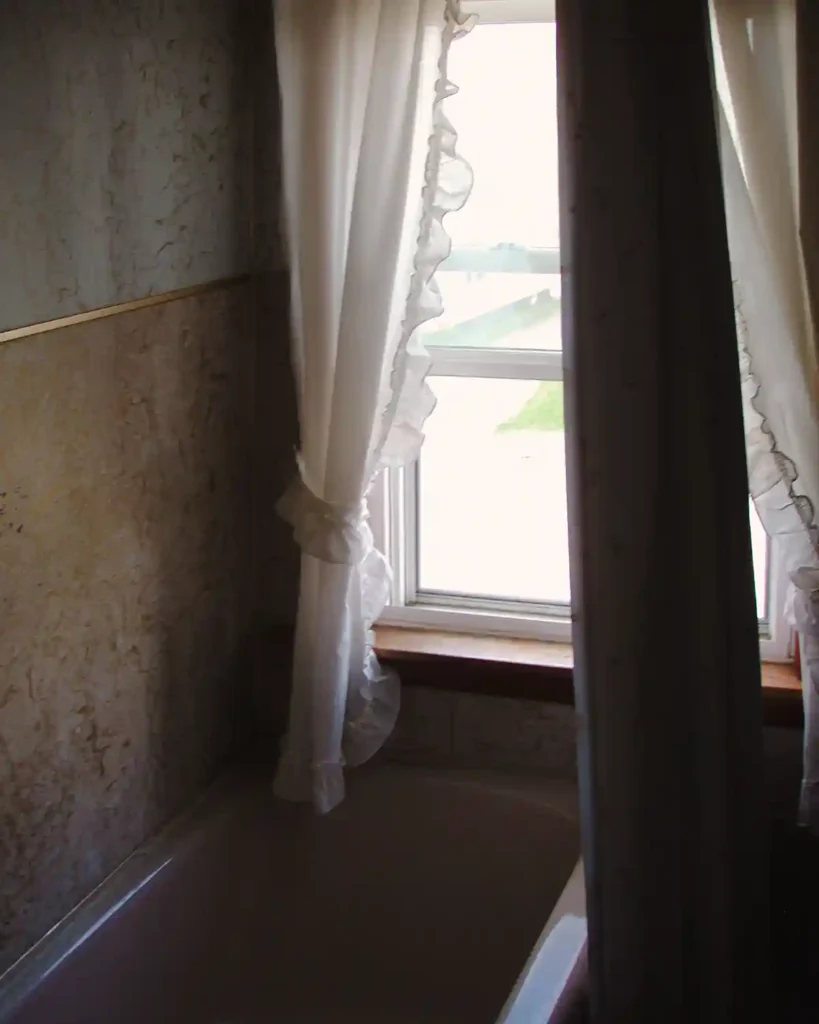 Before tour of a dated bathroom with a pink tub, window at the end of the shower, vinyl walls covered in mold, ruffled curtains, and a vinyl shower curtain hanging