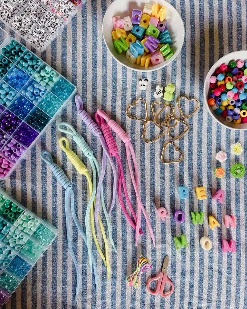 DIY bag charm supplies including paracord, pony beads, gold heart carabiners, a bowl of charms, chunky letter beads, scissors, and elastic string