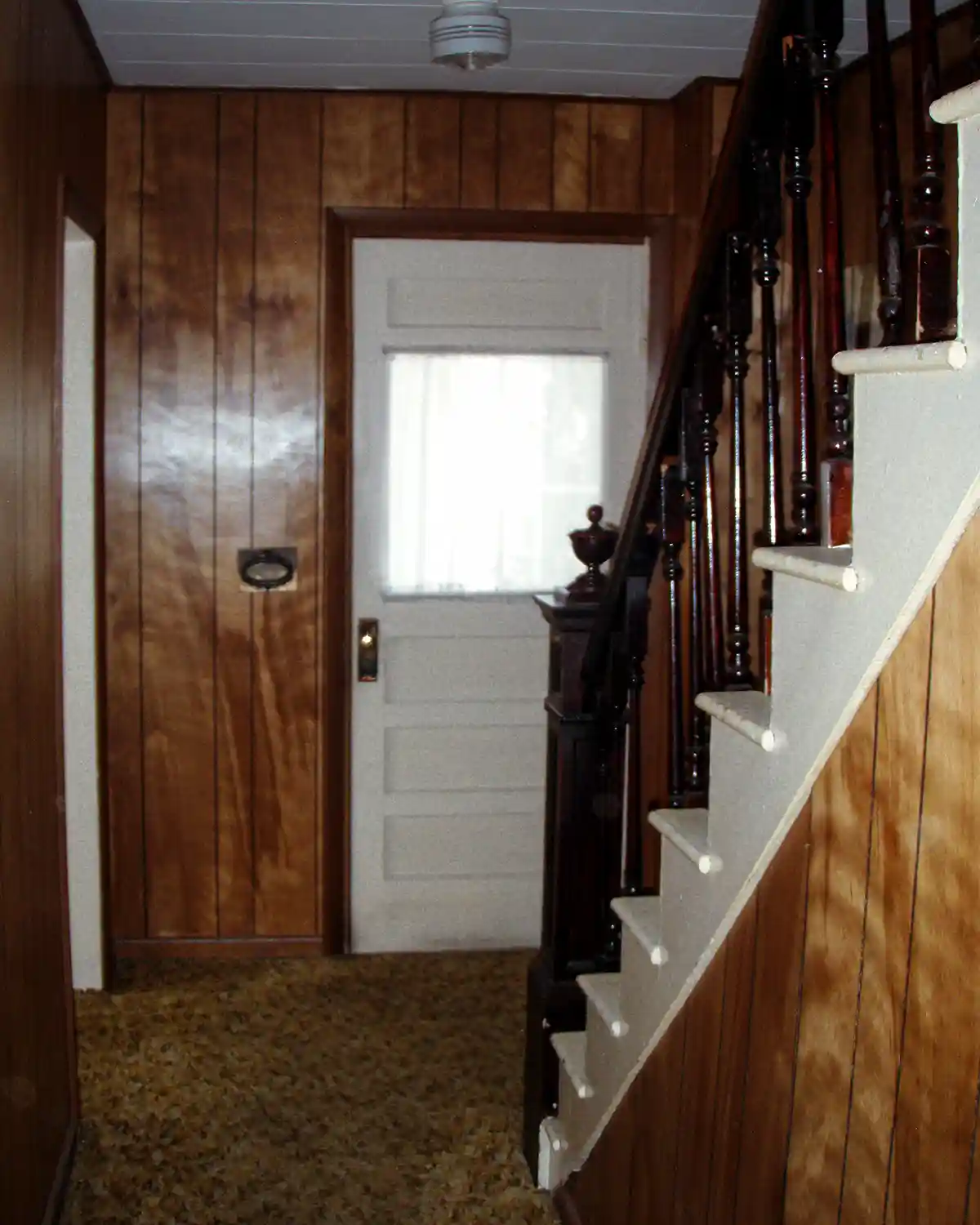 A dark and dated 1970s style hallway with paneled walls, shag carpeting, a front door leading to a porch, and an old staircase with dark stained wood balusters and newell post